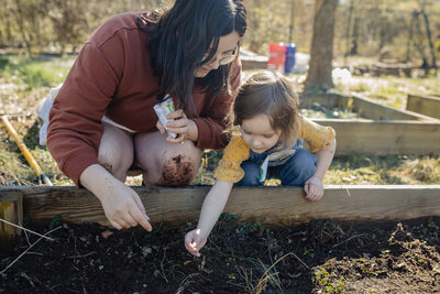 Mom kneels down next to her little girl and shows her how to plant seeds in the garden.