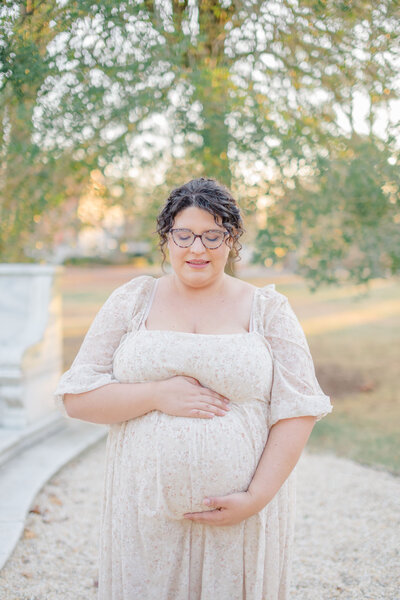 A pregnant mother stands outdoors with her hands gently cradling her belly, looking down with a serene expression. She wears a soft, flowing dress, and the trees in the background create a natural, peaceful setting. The warm lighting highlights her maternal glow. Portrait Studio Stamford CT.