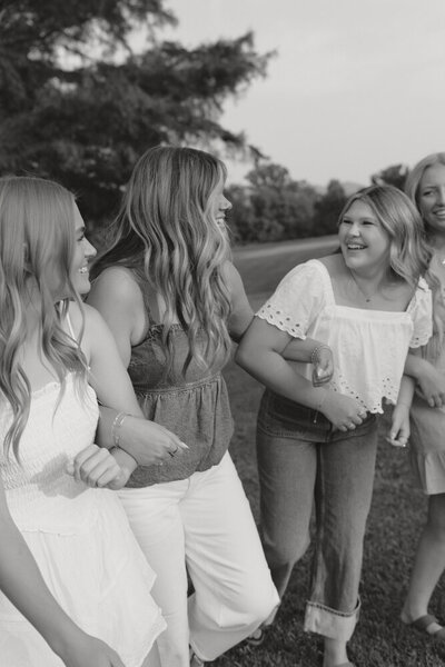 A black and white photo of four senior girls holding hands while they walk and laugh together in a grassy area with trees in Johnson City, TN. 