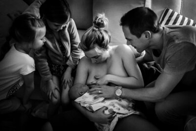 A special shot with the whole family gazing at the just born baby while he is nursing for the very first time. His sisters gaxe on in wonder and the dad supports the mom in getting the baby to latch.