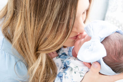 Mom kissing her newborn  wearing a blue bow and flower onesie.