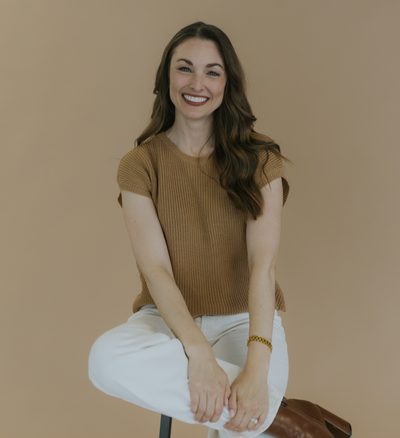 A young woman with brown hair in a camel shirt and white pants sitting on a stool, holding her leg in a relaxed position.