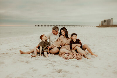 A family of four sitting on a sandy beach near the ocean, with a dock and buildings in the background. The parents are seated in the center, with two children on either side.