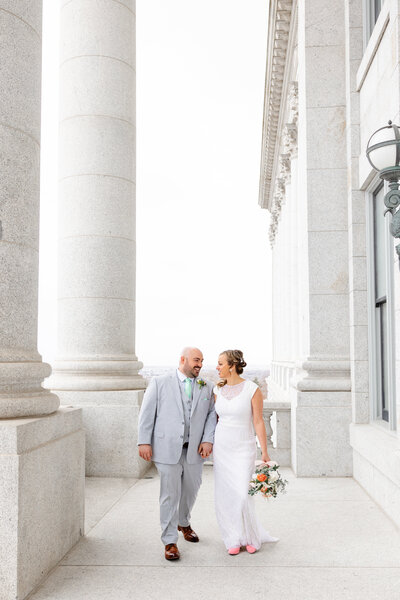 A couple, holding hands, walks as they smile at each other. They are surrounded by towering white columns.