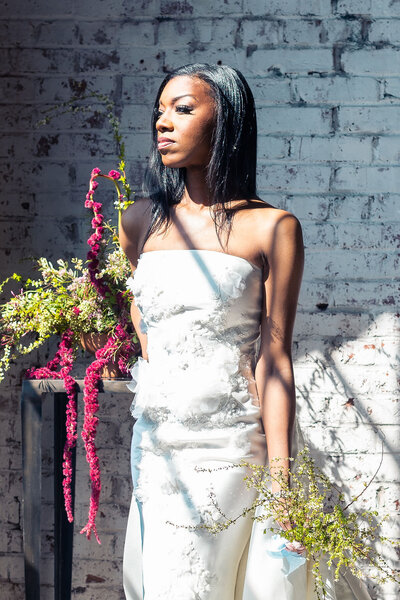 Bride stands by pink and green spirea centerpiece on a wooden table.. She poses against a white brick wall, shadows cascading her dress