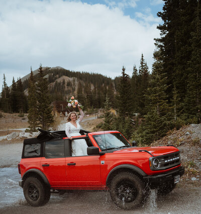 off roading in colorado the couple drove through a large puddle. the bride is standing up outside of the car, which is a bright red off roading bronco. She is holding her flowers above her head and cheering. The groom is driving the car. The bottom of the car is wet from the water.