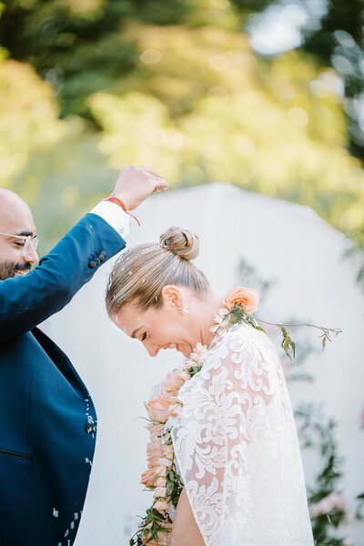 Rhode Island Wedding Photographers | A joyful couple during a wedding ceremony. The groom, in a blue suit, sprinkles petals over the smiling bride, dressed in a lace gown, against a lush green backdrop.