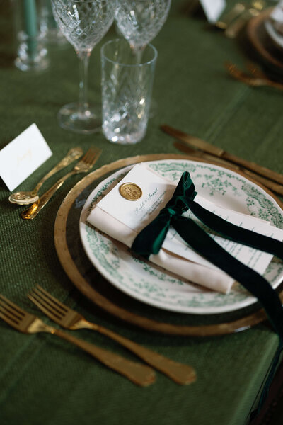 wedding dinner table setting on green linen with gold cutlery and a patterned green charger plate topped with a menu tied with a green bow