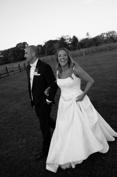 A newlywed couple joyfully walks across an open field, hand in hand, during sunset. The bride, with long, wavy hair and a radiant smile, lifts the skirt of her elegant, textured wedding gown as she walks, exuding pure happiness. The groom, dressed in a sharp black tuxedo with a crisp white dress shirt and bow tie, grins as he leads the way, his boutonniere neatly pinned to his lapel. The rustic setting features a wooden fence and rows of trees in the background, capturing the essence of an intimate long island countryside wedding. The black-and-white tones of the image emphasize the emotion and timelessness of the moment.