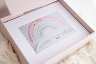 linen collection box of matted portraits with lifestyle newborn portrait of baby in crib and rainbow on wall.