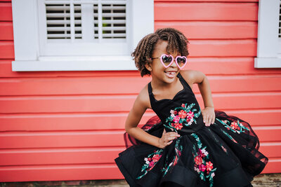African American girl with curly brown hair and pink hearts glasses with hands on hips wearing a black floral dress in fells point Baltimore Maryland