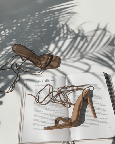 A pair of high-heeled sandals resting on an open fashion magazine with sunlight casting shadows, adding an elegant touch.