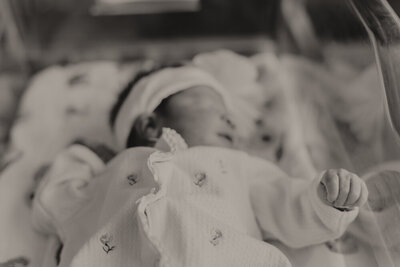 A sweet black and white photo of a newborn girl laying in her hospital bassinet with pajamas and a headband on