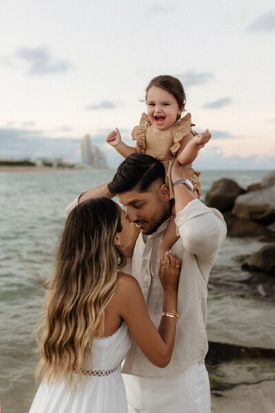 Family portrait on a Miami beach, featuring parents holding their baby with ocean waves in the background