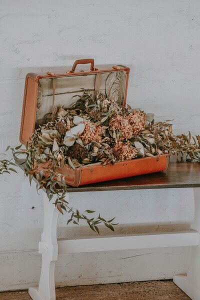 Scénographie proposée par une décoratrice de mariage. Valise vintage marron posée sur une table remplie de vegetal.
