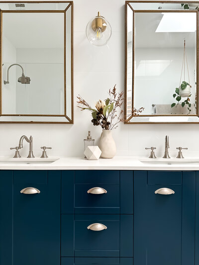 Modern bathroom with double sinks, navy cabinets, and brass mirrors crafted by a Newcastle Interior Designer. A vase with dried flowers adds a touch of elegance on the counter.