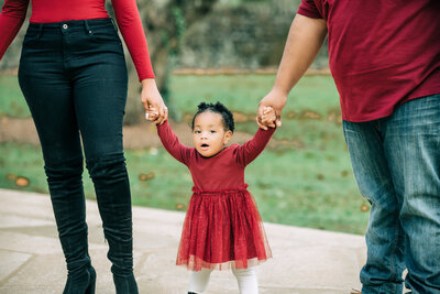 Little girl about 2 years old in red frilly dress holding both parents hand trying to walk.