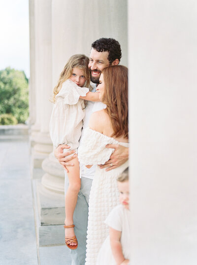 Dad embraces daughter and wife while standing next to stone columns
