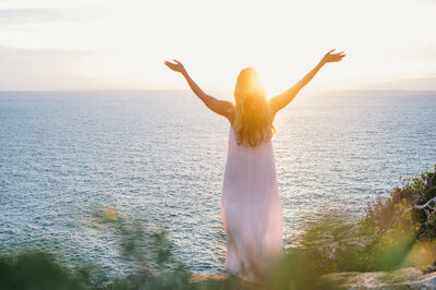 A woman in a flowing white dress raises her arms toward the sunrise while standing on a cliff overlooking the ocean.