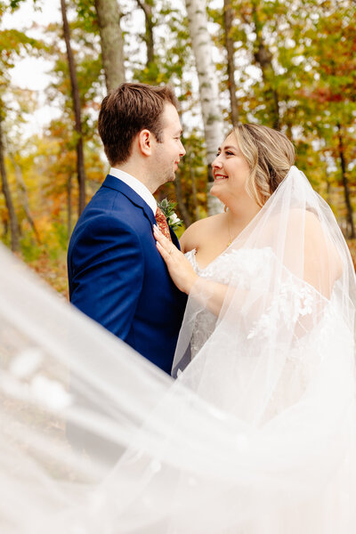 A bride and groom stand close in a wooded area with autumn foliage, joyfully captured by a Minnesota photographer. The bride, in her veil and lace dress, smiles at the groom in his blue suit, both surrounded by colorful leaves—a perfect moment of wedding photography bliss.