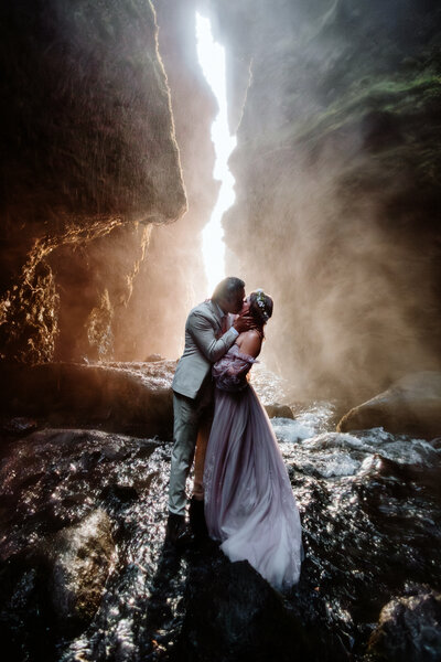 Fairytale newly wed couple is kissing in a cave with a waterfall and sparkly light coming through the cave in Iceland on their elopement day