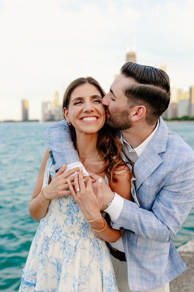 Man kisses his fiance's cheek on the North Ave Beach with the Chicago skyline behind them