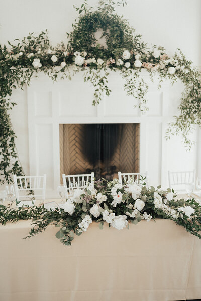 Fresh greenery florals drape over white fireplace behind bride and groom seats at reception