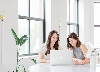 two women working at a Macbook laptop, smiling a looking down a the screen together
