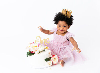 Baby girl in a white lace romper and floral crown poses for her first birthday cake smash. She is covered with icing on her hands, face, and knees. Cake is smashed. Baby is smiling at the camera. Captured in Brooklyn, NY.