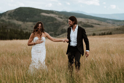 Bride and groom walk hand-in-hand through golden field, laughing during mountain wedding near Breckenridge, CO.