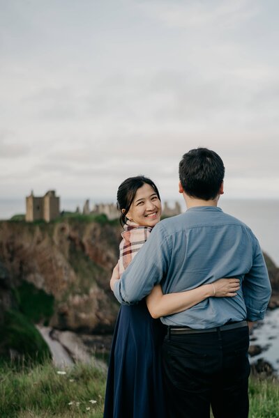 A close view of an engaged couple standing side by side with their bodies pressed together on a cliff beside Dunnottar Castle in Aberdeenshire.