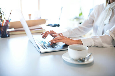 A person in a white blouse types on a laptop at a white desk. There's a cup of coffee on a saucer nearby, along with stacks of papers and a cup holding pens in the background. The scene is brightly lit, suggesting daylight.
