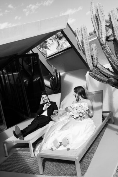 A black-and-white photo of a bride and groom relaxing on lounge chairs at a mid-century modern venue in Palm Springs, with desert landscaping in the background.