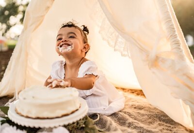 Smiling baby girl sitting under a cream teepee next to a birthday cake during a whimsical outdoor cake smash session in Charleston, SC.