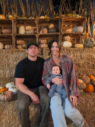 A man and woman sit on a hay bale with their baby, enjoying a family moment at the pumpkin patch.