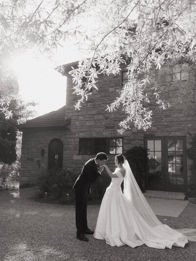 Groom kissing bride’s hand during outdoor wedding, black-and-white photo.