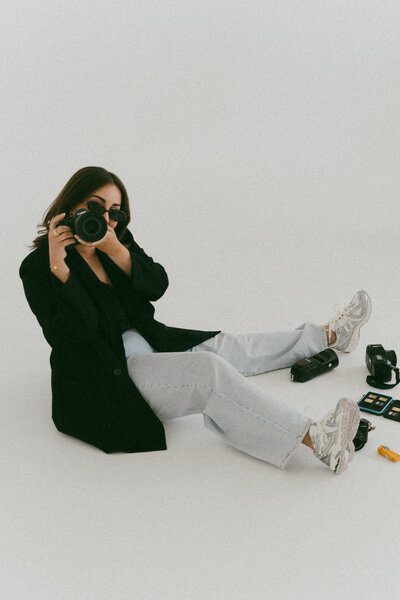 Female photographer sitting on the studio floor in light denim and black blazer, playfully aiming her camera toward the lens.