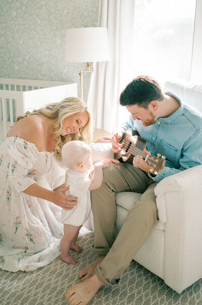 a father plays guitar for his family, while his toddler and wife smile listening to him play