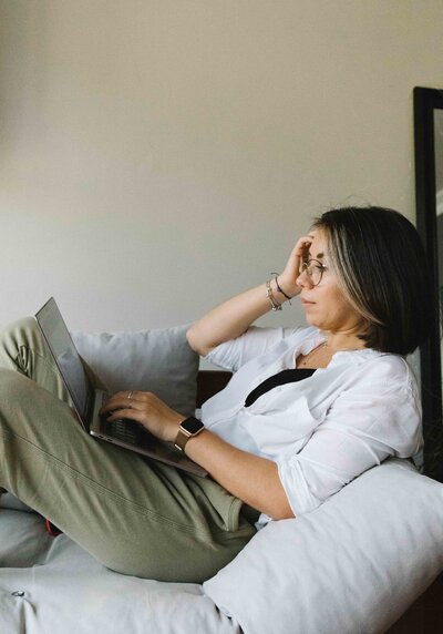 A woman is shown in profile, reclining against the arm of a couch, with her right hand resting on her forehead. She is typing on a laptop with her left hand, a focused expression on her face.