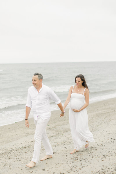 Parents-to-be walk holding hands along Sherwood Island beach during maternity portrait session