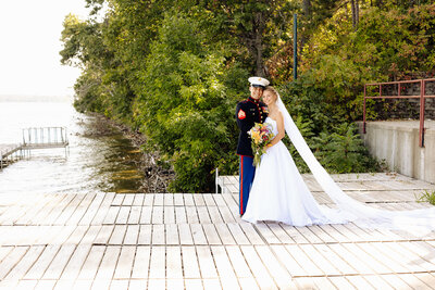 A couple stands on a wooden dock by a lake, captured beautifully by Minnesota photographer Amber Bauer. The groom is in a military dress uniform, and the bride wears a long white gown with a veil. Smiling, they hold a bouquet of flowers as trees line the water's edge under the bright sky.