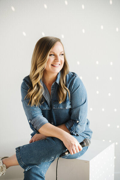 A photo of Krista Marie, sitting on a white block, holding a microphone and smiling and looking away from the camera