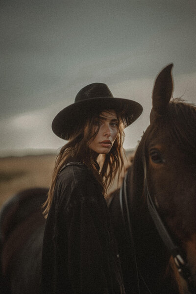 A woman wearing a black hat and dark clothing stands beside a brown horse outdoors under a cloudy sky, embodying the elegance and detail found in Sweet Pea Ink Creative's brand design.