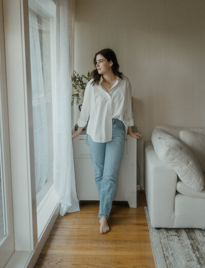 Woman wearing a white button down and blue jeans leaning back against a dresser and looking out a window