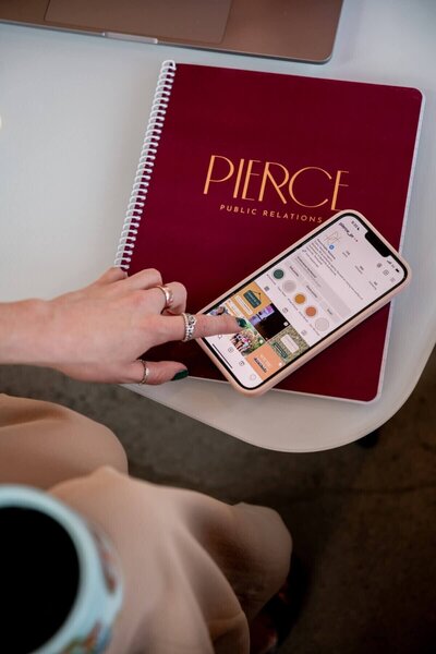 Hand scrolling through Pierce PR’s Instagram profile on a smartphone, placed on top of a branded maroon notebook.