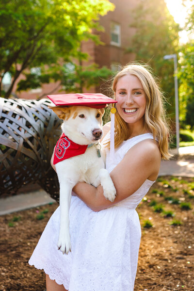 NC State graduate in white dress smiling while holding her dog wearing a red bandana and graduation cap.