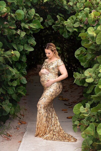 Beach maternity portrait in gold dress under greenery arch in West Palm Beach.