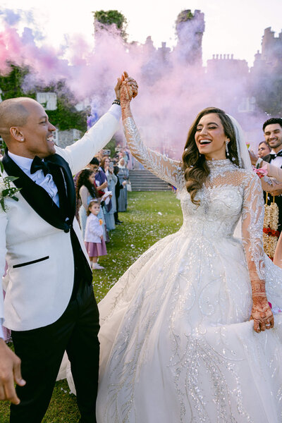 Bride & Groom hold hands together high in the sky with pink & blue smoke bombs going off in the backround to celebrate their wedding at Luttrellstown Castle