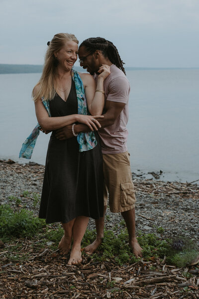 A photo of a couple at the beach in Ithaca, NY. They are on the shore, with the bright blue sky as their backdrop. The mand is standing just behind the woman and wrapping his arms around her to embrace her. Photo by Dash and Boheme Photography. 