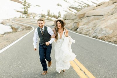 An adventurous couple run up the road surround by mountains on their mountain wedding day. 